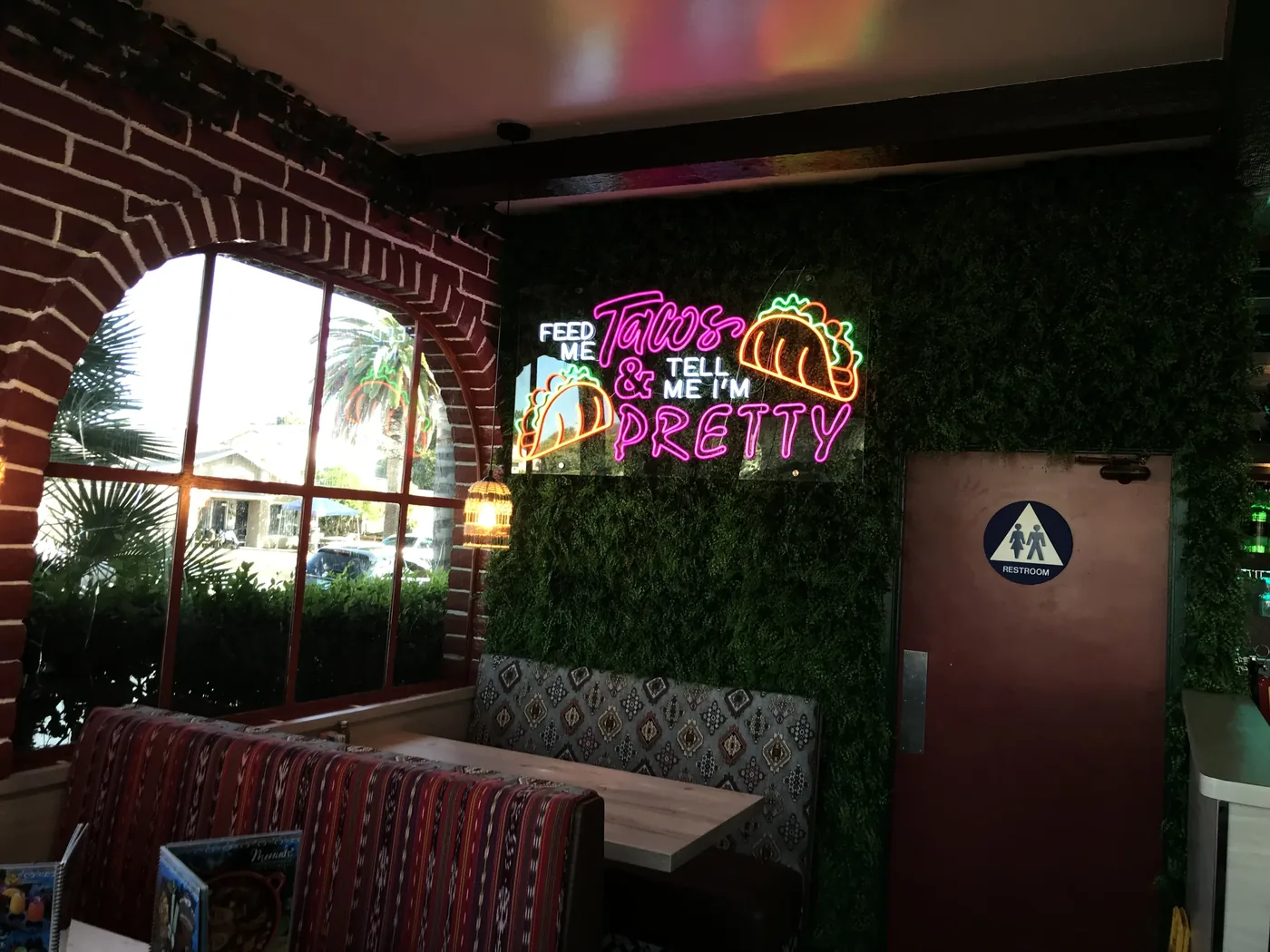 Feed Me Tacos neon sign on a greenery wall inside a restaurant