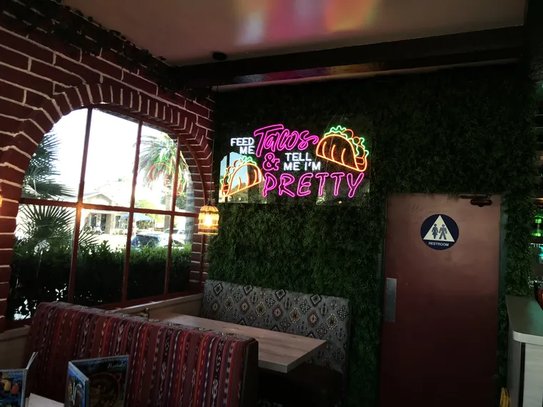 Feed Me Tacos neon sign on a greenery wall inside a restaurant